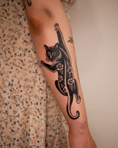 Chat noir qui grimpe, entouré de fleurs, tatoué sur un bras